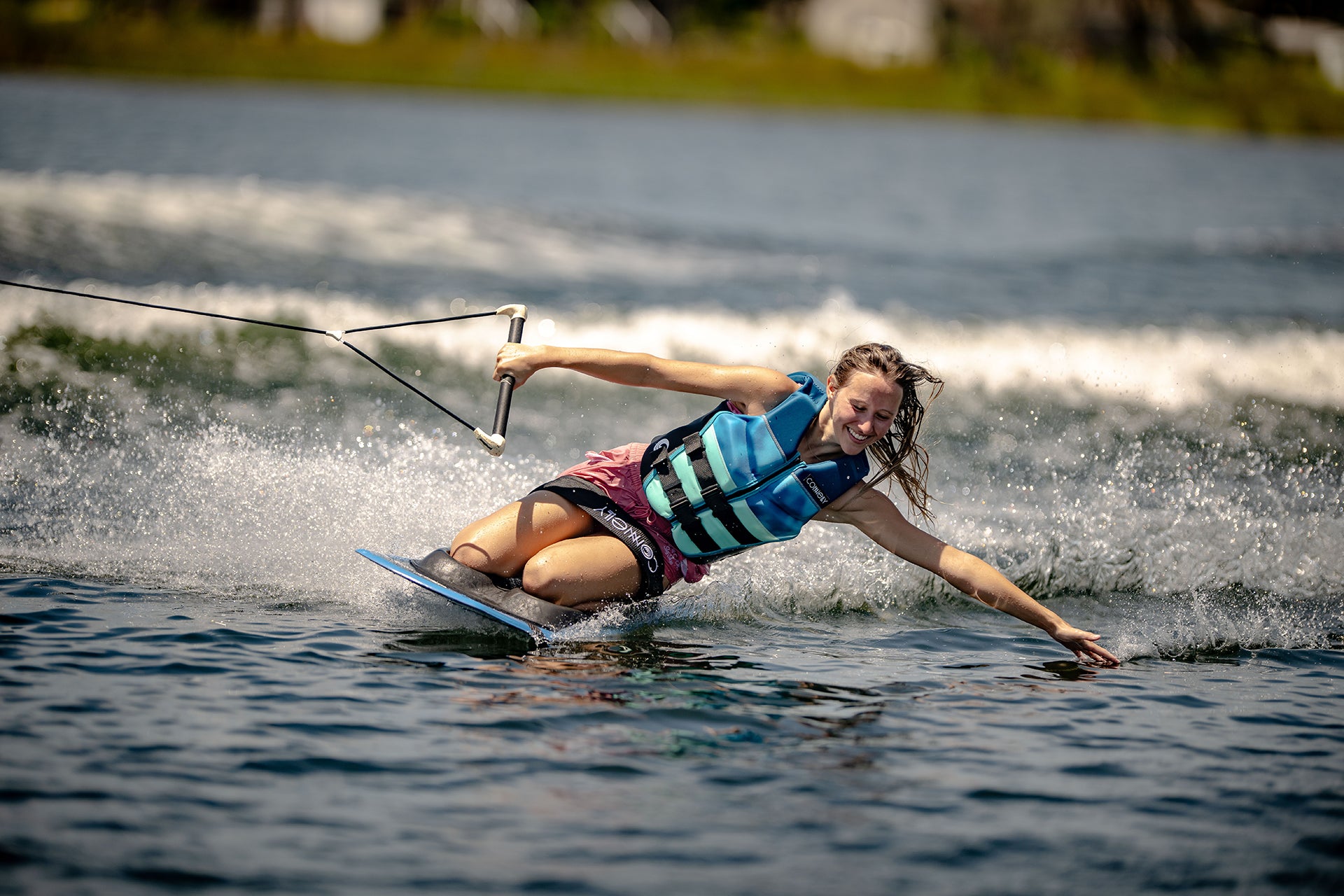 Person wakeboarding, wearing a blue life jacket, skimming the water with one hand.