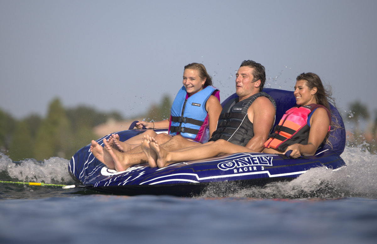 Three riders in life jackets gripping the handles as the Racer 3 towable tube glides over the water, splashing around them.