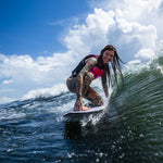 A female surfer in a pink life vest carves a wave on the Seer wakesurf board, crouching with a smile.