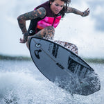 A female surfer launches into the air on the Seer wakesurf board, showcasing its sleek gray base with black accents.