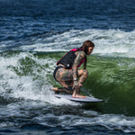 A female surfer crouches low while riding the Seer wakesurf board, maintaining balance on a green wave.