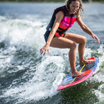 Woman in a pink and black life vest wakesurfing on the SPARK board, smiling with arms out for balance.