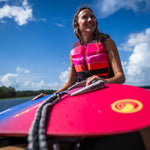Woman in a pink and black life vest holding the SPARK wakesurf board on a boat with a lake in the background.