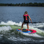 Rear view of a woman wakesurfing on the SPARK board, creating a foamy white wave trail on the lake.