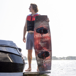 Rider holding the 2024 Standard wakeboard, featuring a red geometric design and black bindings while standing on a boat.
