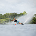A male water skier wearing a life vest carves through the water on a Connelly C1 ski, creating a spray near a red buoy.