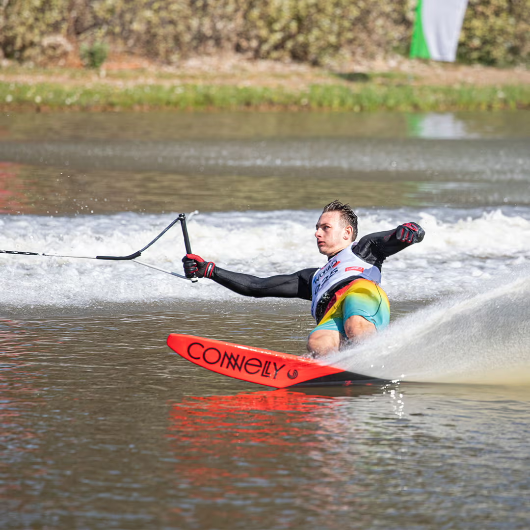 Person water skiing on one ski, creating a spray of water, with trees in the background.