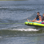 Two riders sitting upright, gliding across the water on the Fun 2, a bright blue and yellow inflatable towable tube.