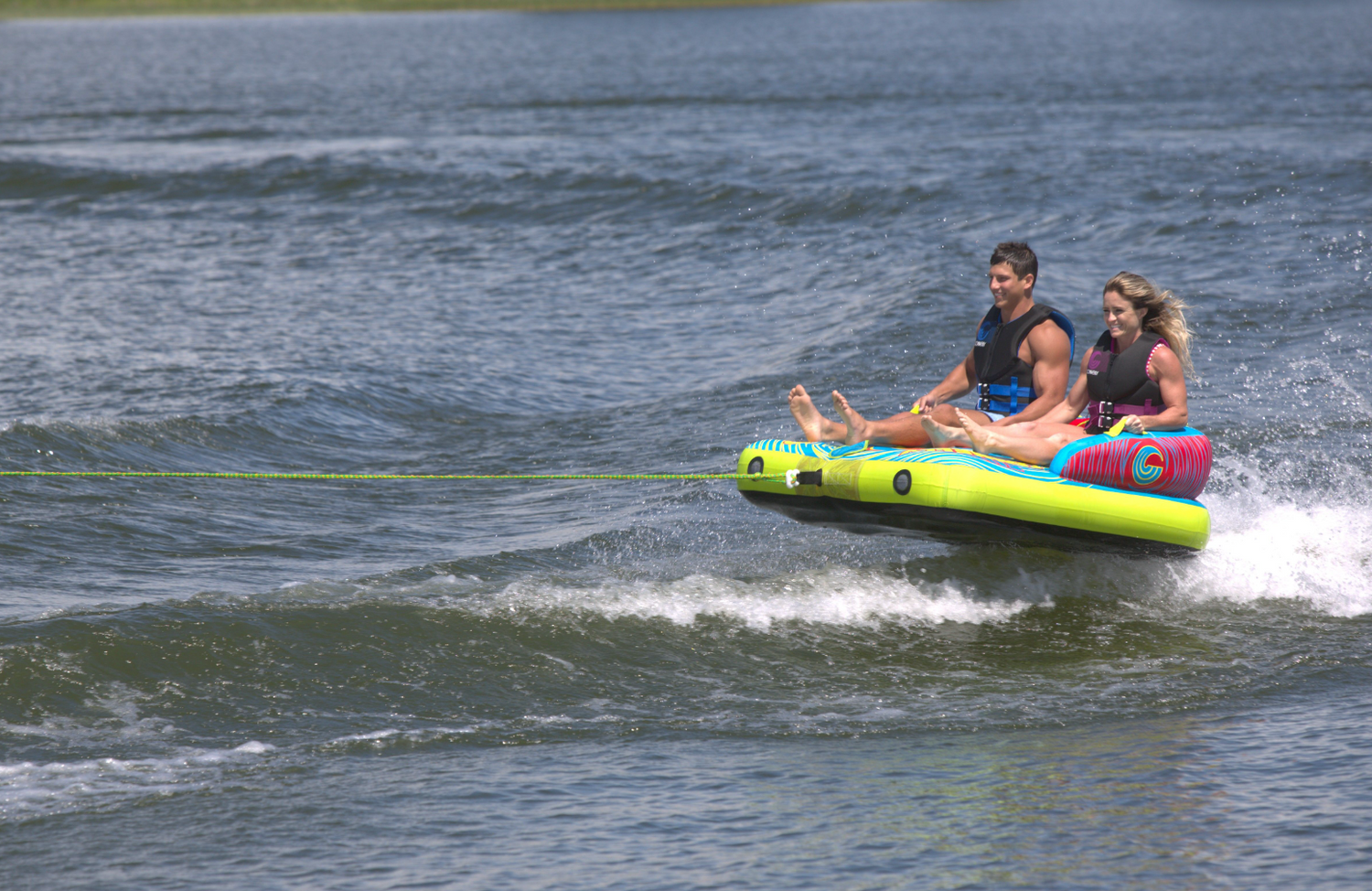Two riders sitting upright, gliding across the water on the Fun 2, a bright blue and yellow inflatable towable tube.