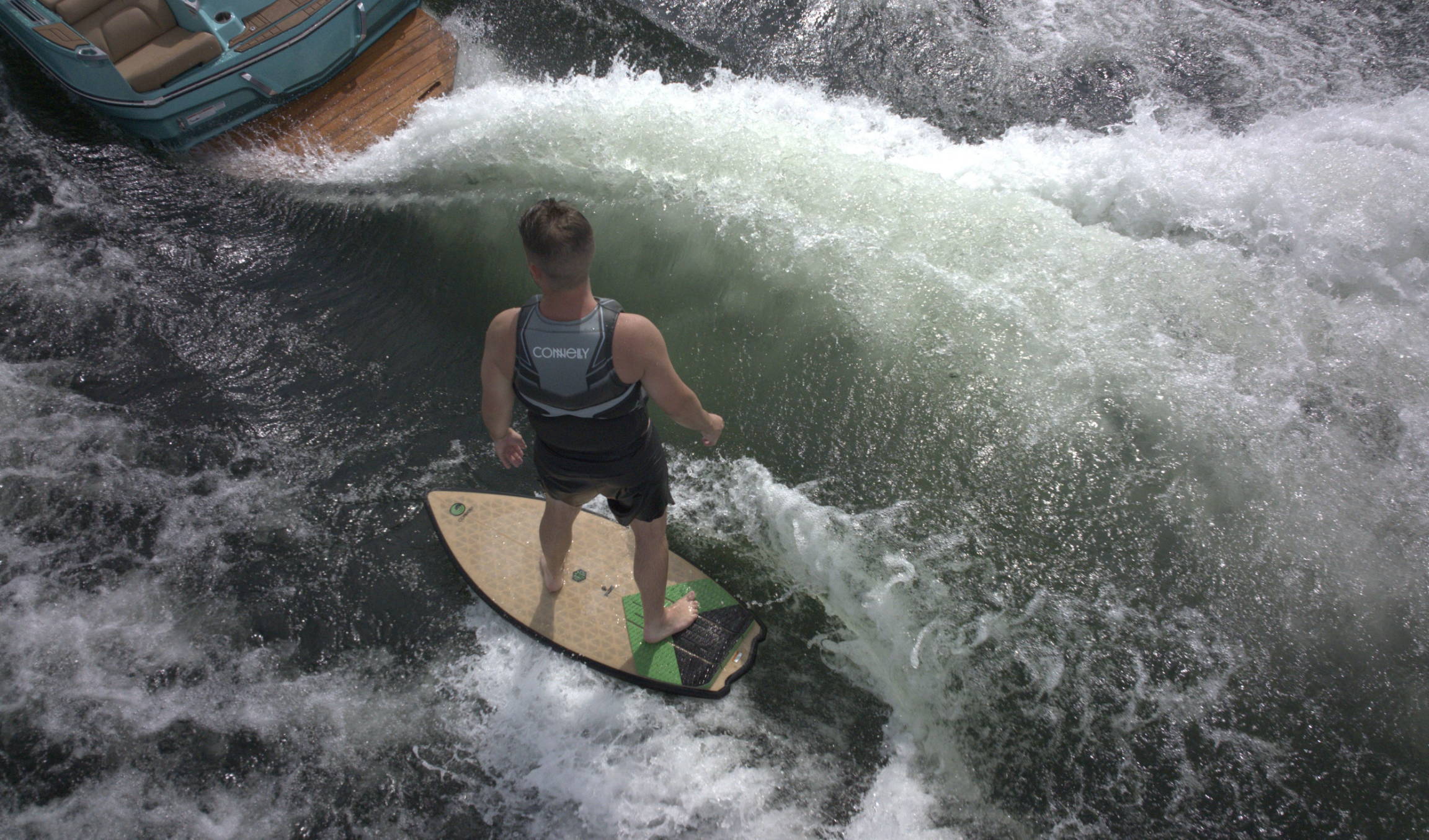 A wakesurfer riding a wave behind a teal boat, wearing a Connelly life vest and balancing on a tan and green surfboard.