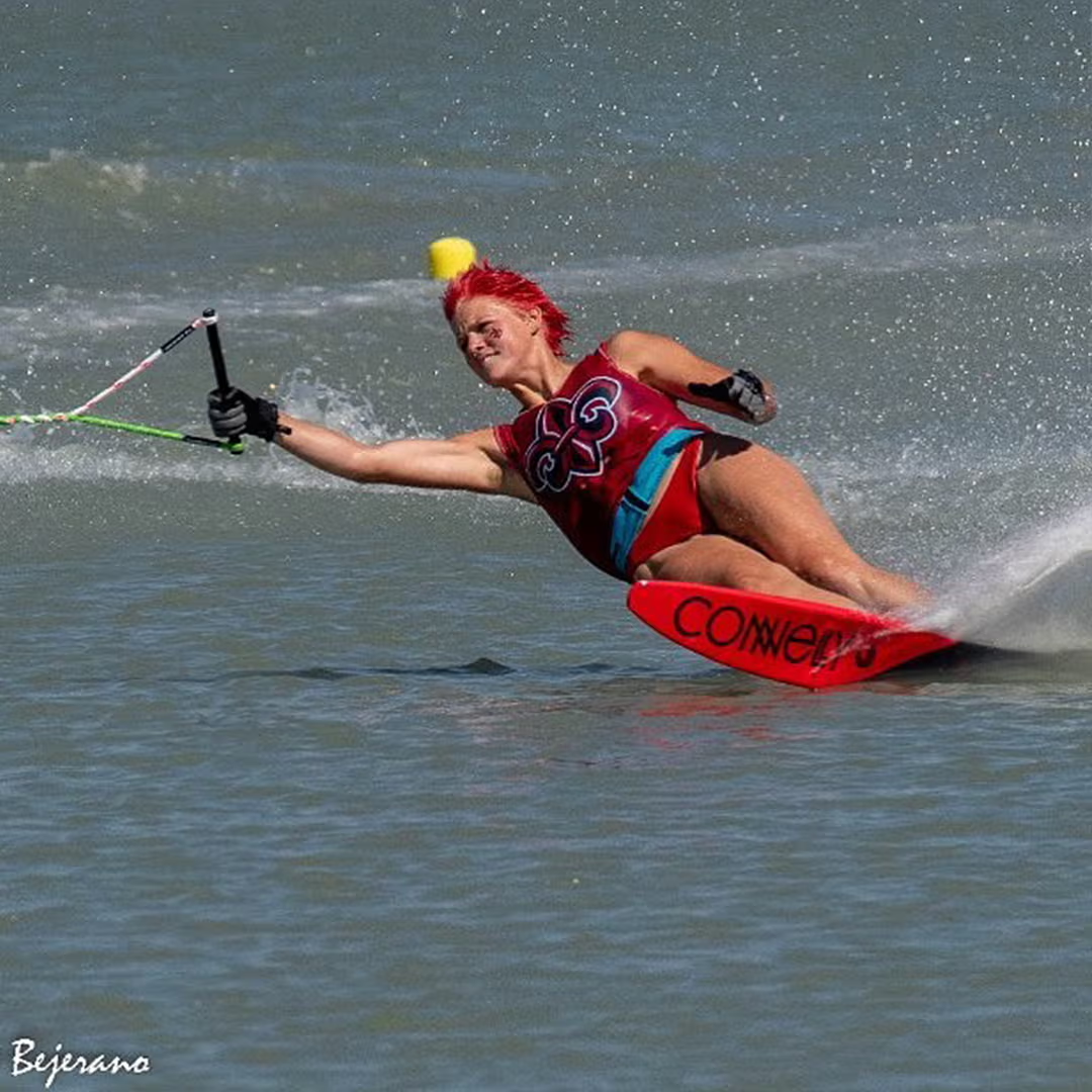 Person with red hair water skiing at high speed, leaning sharply with spray around.