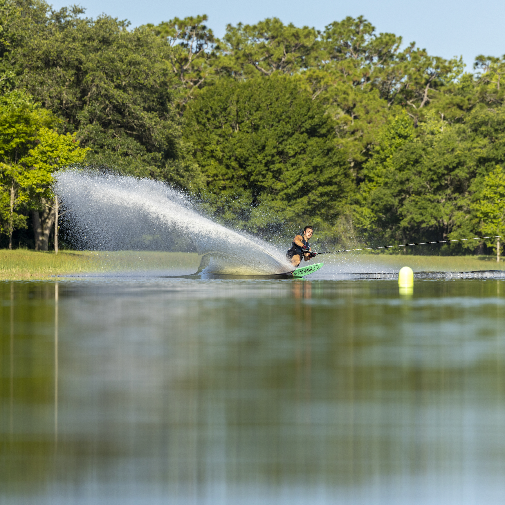Person waterskiing on a lake, creating a large spray of water, with trees in the background.