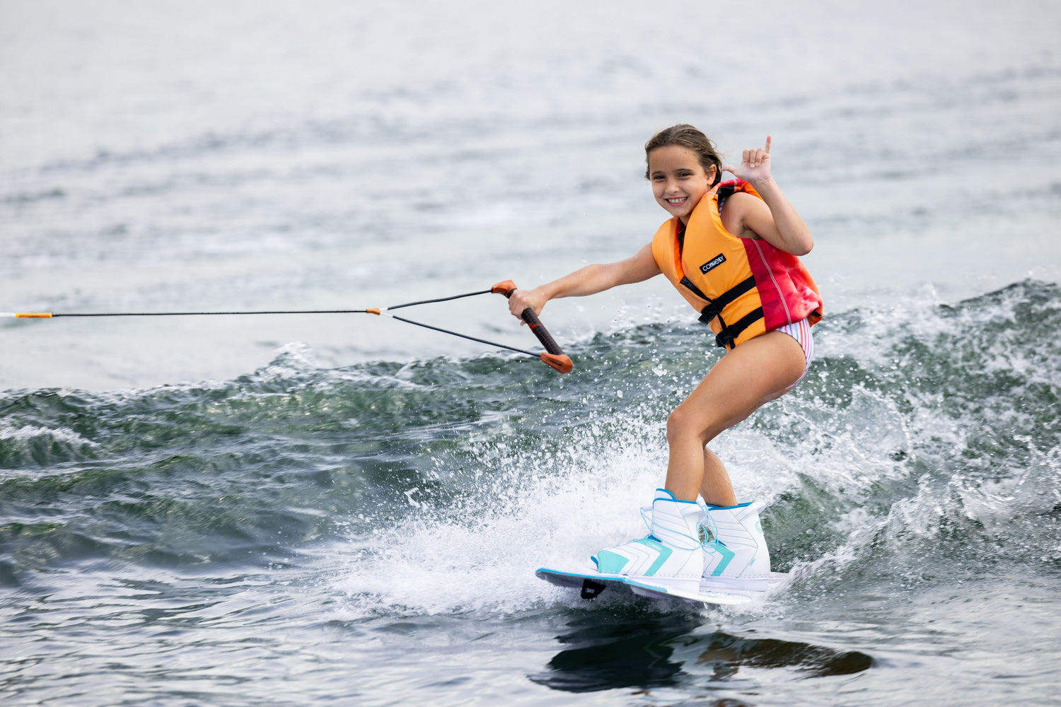 Youth wearing a Bella life jacket wakeboarding on a lake, making a hand gesture.
