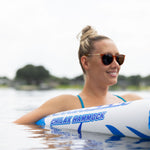 A woman wearing sunglasses and a blue swimsuit floats in the water on the Chilax Hammock, smiling while partially submerged.