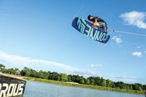 The 2024 Groove wakeboard in mid-air, showcasing its bold blue and teal graphics with dynamic water spray.