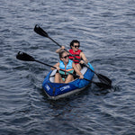 A woman and a man paddle the Nautic 11.5 inflatable kayak on a lake, both wearing life jackets and sunglasses.