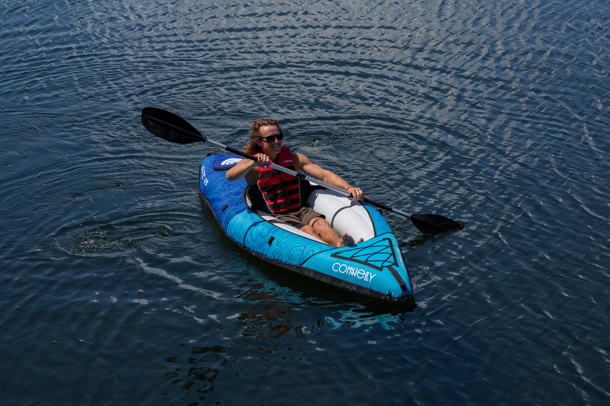 A man in a red life jacket paddles the Nautic 9.5 inflatable kayak on a calm lake.