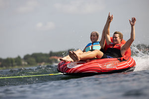 Two riders in life jackets enjoying the Racer 2 towable tube, speeding over the water with splashes around them.