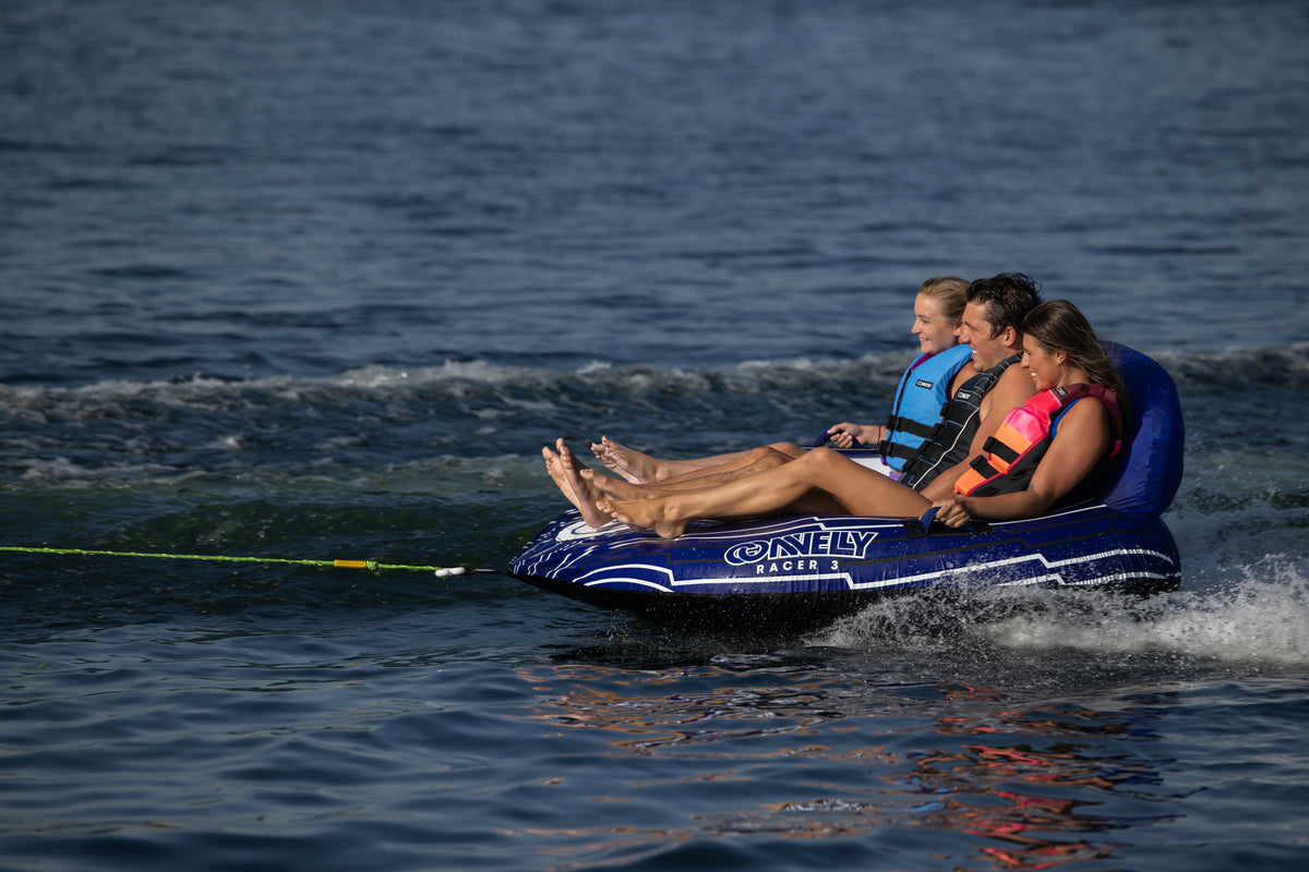 Three people riding the Racer 3 towable tube, leaning back as they speed across the water with a tow rope attached.