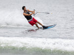 A male water skier in red shorts and a black vest carves across the water on the Shortline ski.