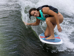 A female wakesurfer riding the Tracer Skim on a wave, leaning into the turn with a smile.